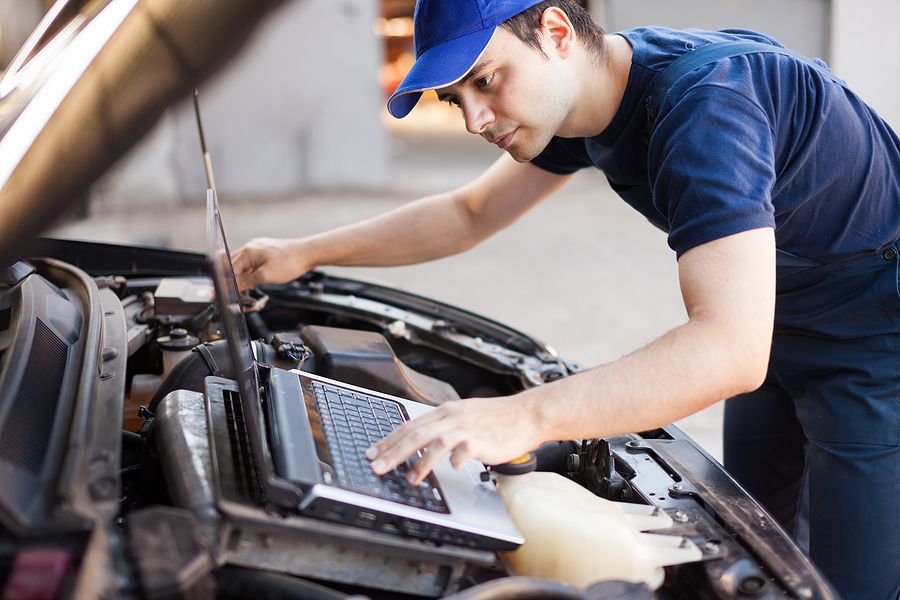 A man is working on a car with a laptop under the hood.