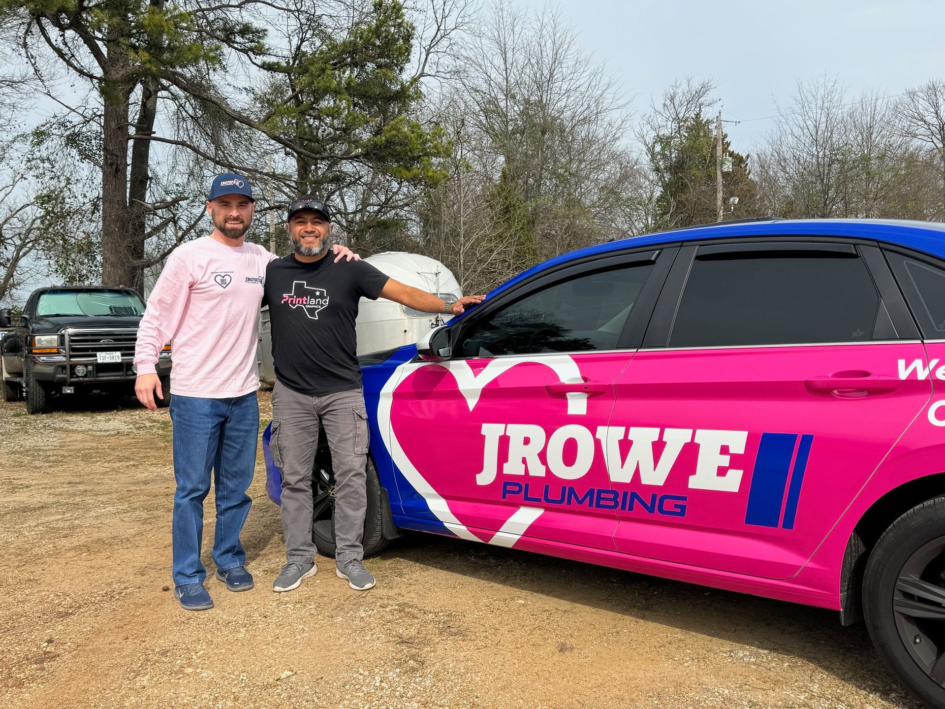 Two men are standing next to a pink and blue jrowe plumbing car.