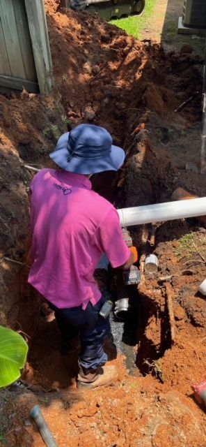 A man in a pink shirt and blue hat is working on a pipe in the dirt.