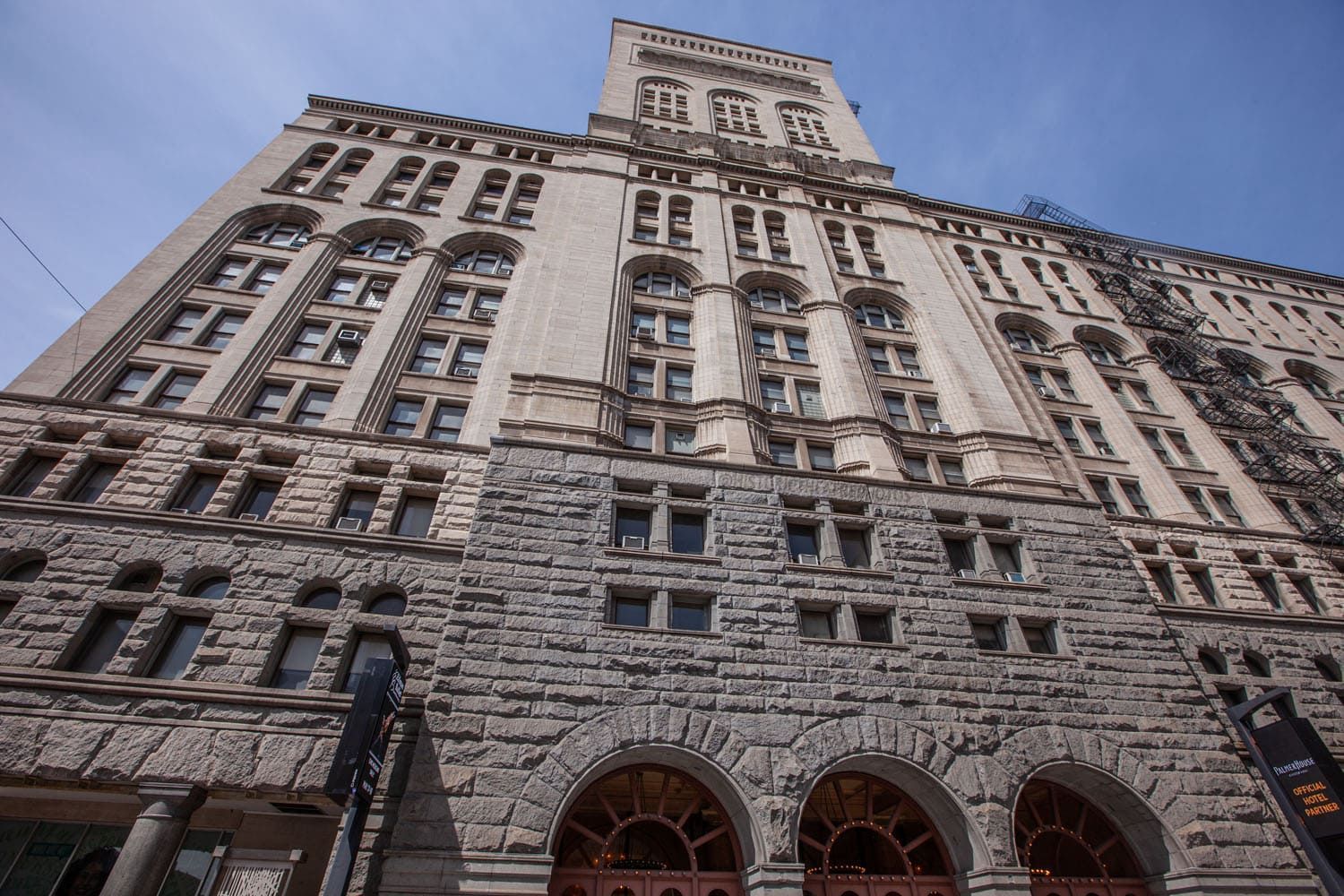 A low-angle view of a grand stone building with arched windows and a prominent central tower against a clear blue sky.