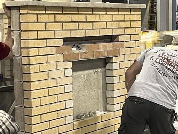 Live bricklaying demonstration at the lintel station during a masonry workshop, showing full mortar joints, proper brick coursing, and installation sequencing over a steel lintel support embedded in the masonry wall.