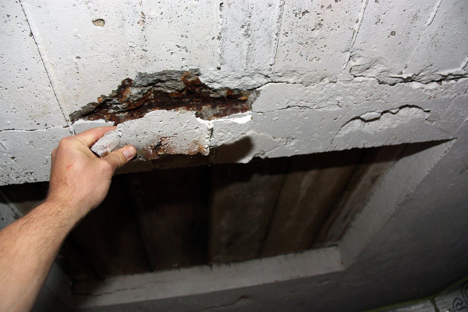 Delaminated concrete slab with a hand lifting a loose section, exposing rusted reinforcing steel caused by moisture intrusion and corrosion—an early warning sign of structural deterioration requiring concrete repair.