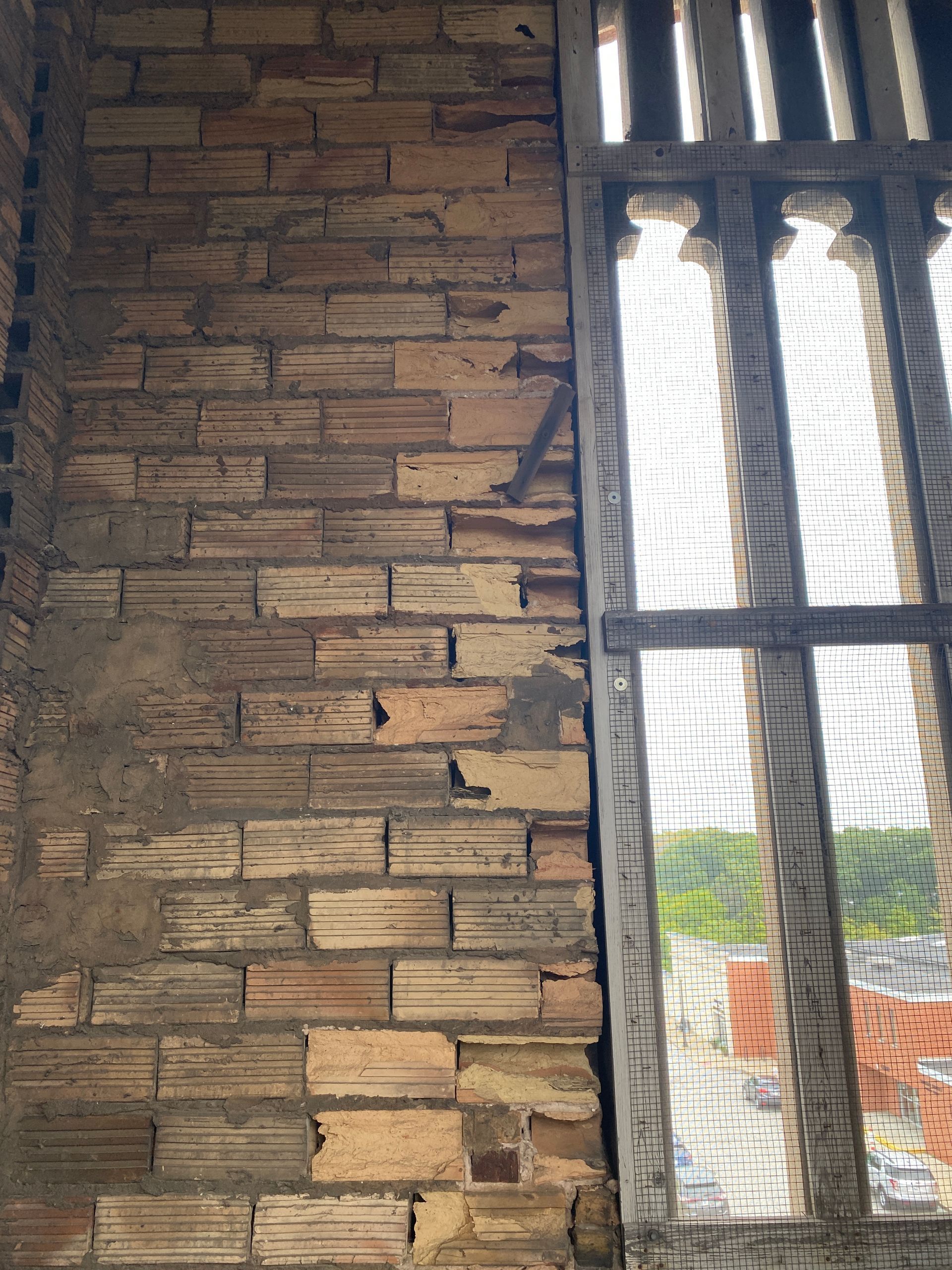 Close-up of weathered brick wall showing freeze-thaw damage, with gaps and uneven surfaces, adjacent to a decorative window in an old building