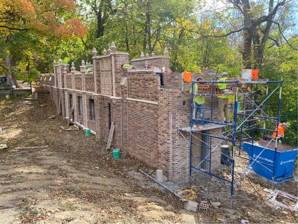 RestoreWorks team members working on scaffolded sections of a detailed brick structure during tunnel brick replacement at the Indiana School for the Blind. The surrounding area features a wooded landscape with autumn leaves. Workers are carefully rebuilding portions of the brick façade to restore the structure's integrity and maintain its historic design.