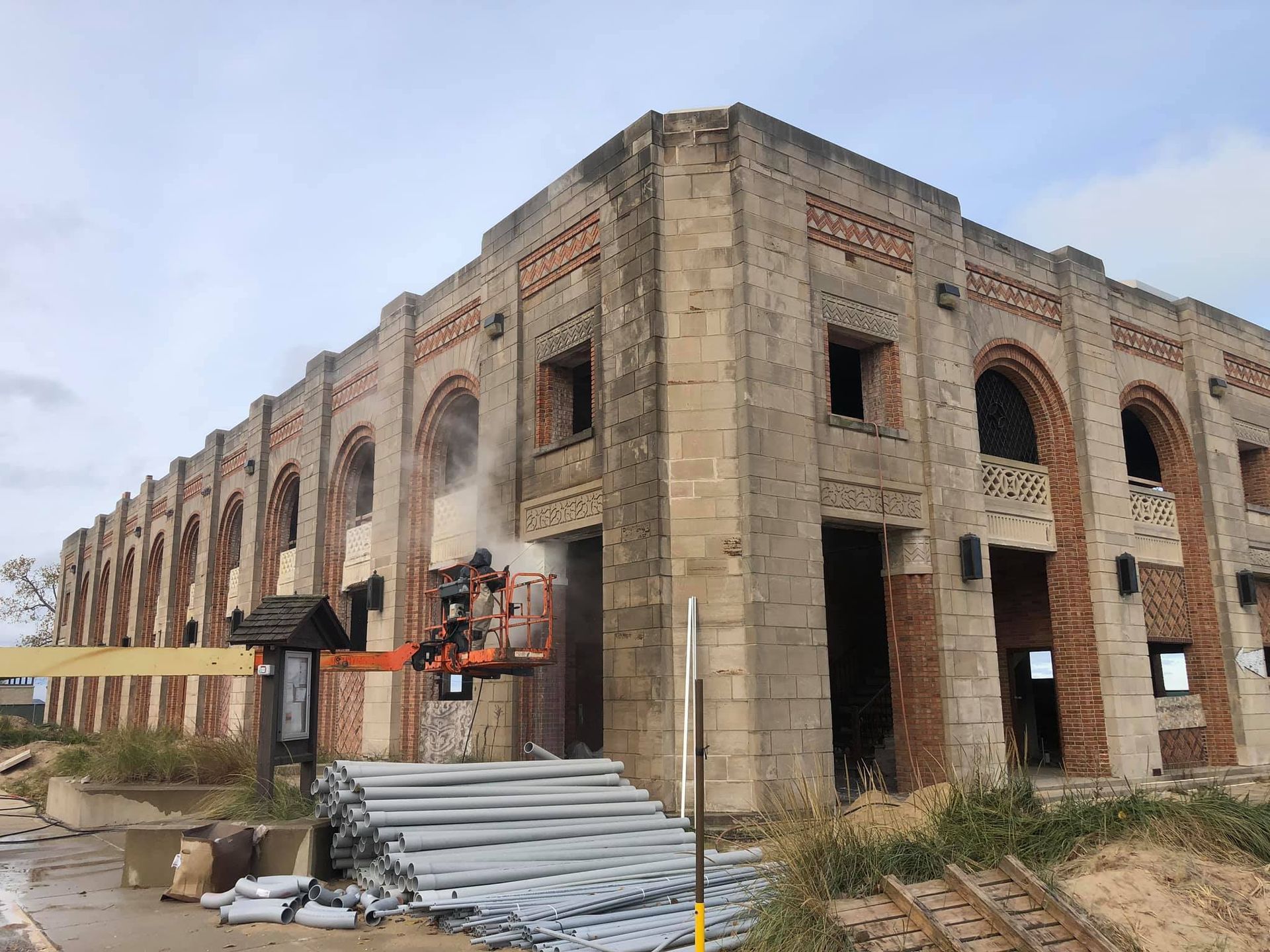 A large historic building undergoing masonry restoration at Indiana Dunes National Park. Workers on an orange lift are repairing brick and stone details on the façade, which features arched windows, intricate stone carvings, and a mix of brick and limestone materials. Construction materials, including pipes and wooden planks, are stacked nearby, with sand dunes and grasses visible in the background.