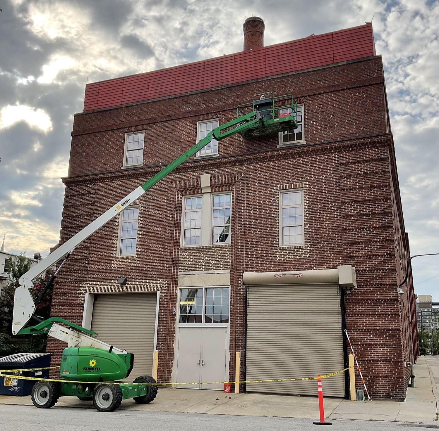 Green lift basket working on a red-brick building with roll-up doors under a cloudy sky