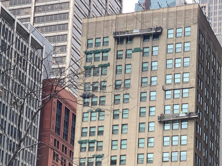 High-rise buildings in a city, with workers on exterior scaffolding platforms performing maintenance on a beige facade.