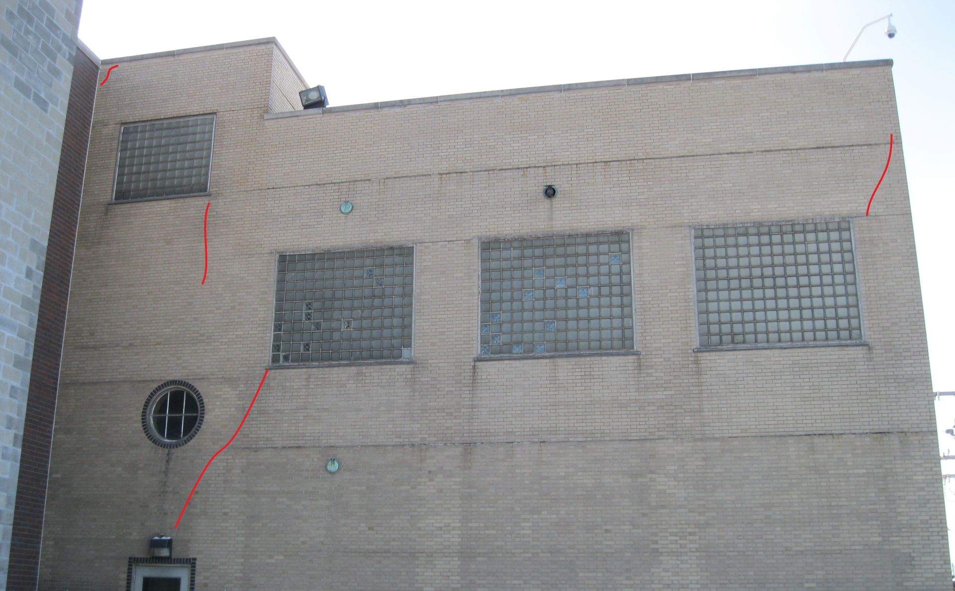 A beige brick building marked with red lines highlighting areas of cracked bricks identified during an inspection. The building features glass block windows and a round decorative window on the lower section.