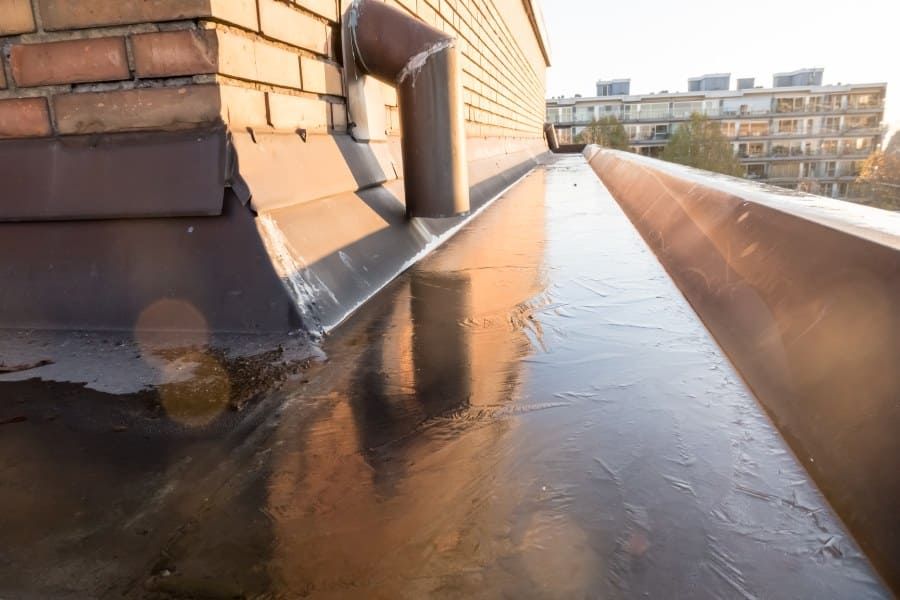 Flat rooftop with a thin layer of ice forming near a brick parapet wall and metal drainage system.