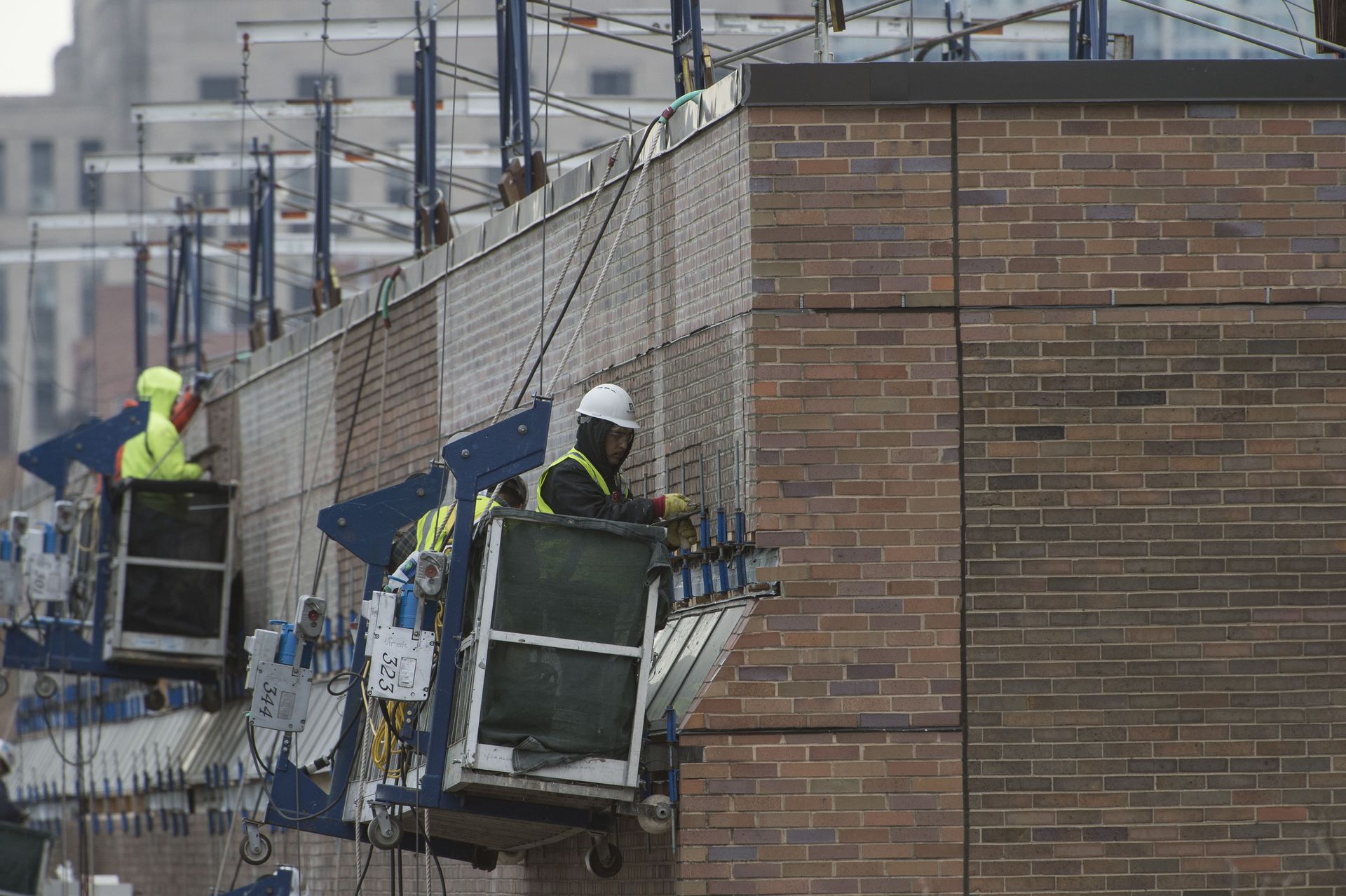 RestoreWorks masonry restoration workers on suspended scaffolding repairing a brick facade at UIC Chicago. One worker in a hard hat and high-visibility vest is carefully replacing damaged bricks, while others perform restoration tasks along the building exterior.