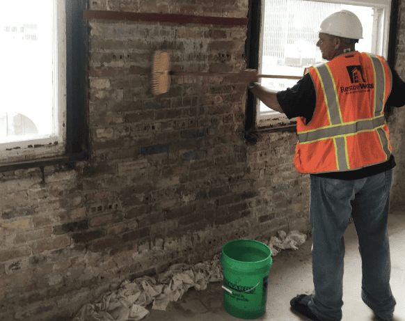 RestoreWorks worker cleaning an interior brick wall with a scrub brush to expose cracks and mortar deficiencies prior to masonry repair.