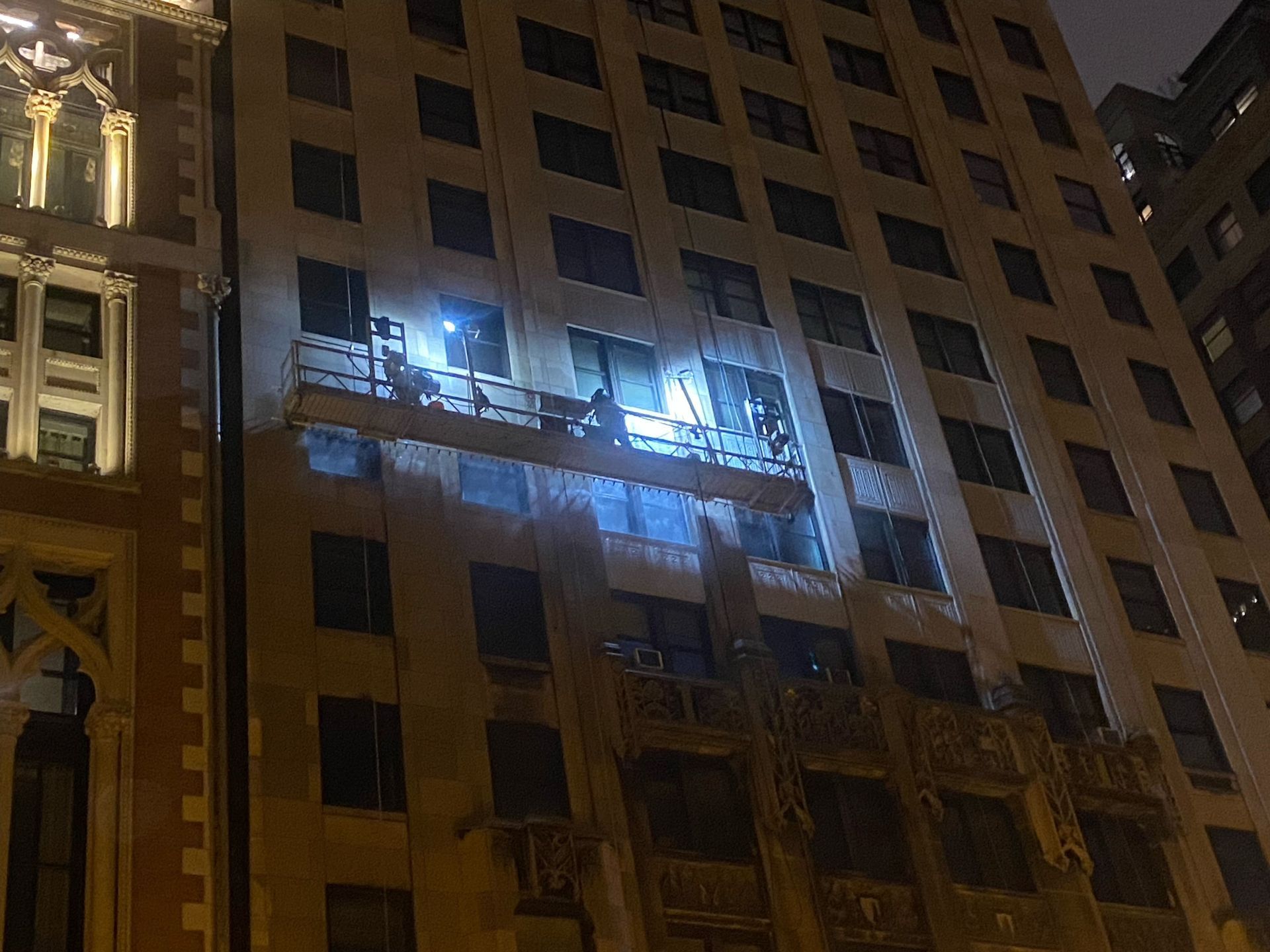 Nighttime view of a tall building with bright lights illuminating a middle-floor balcony or scaffolding.