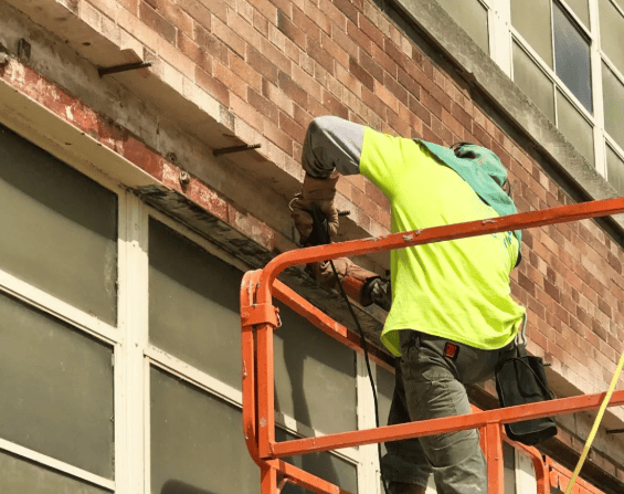 A worker in a high-visibility yellow shirt on a cherry picker uses a power tool to cut into a brick wall.