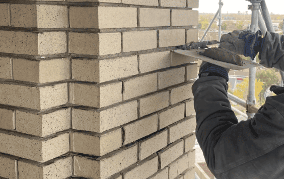 A worker on scaffolding uses a trowel to apply mortar between light-colored bricks on a masonry wall.