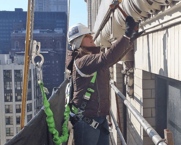 A worker wearing a hard hat and safety harness uses a hammer to perform masonry maintenance on a high-rise building facade.