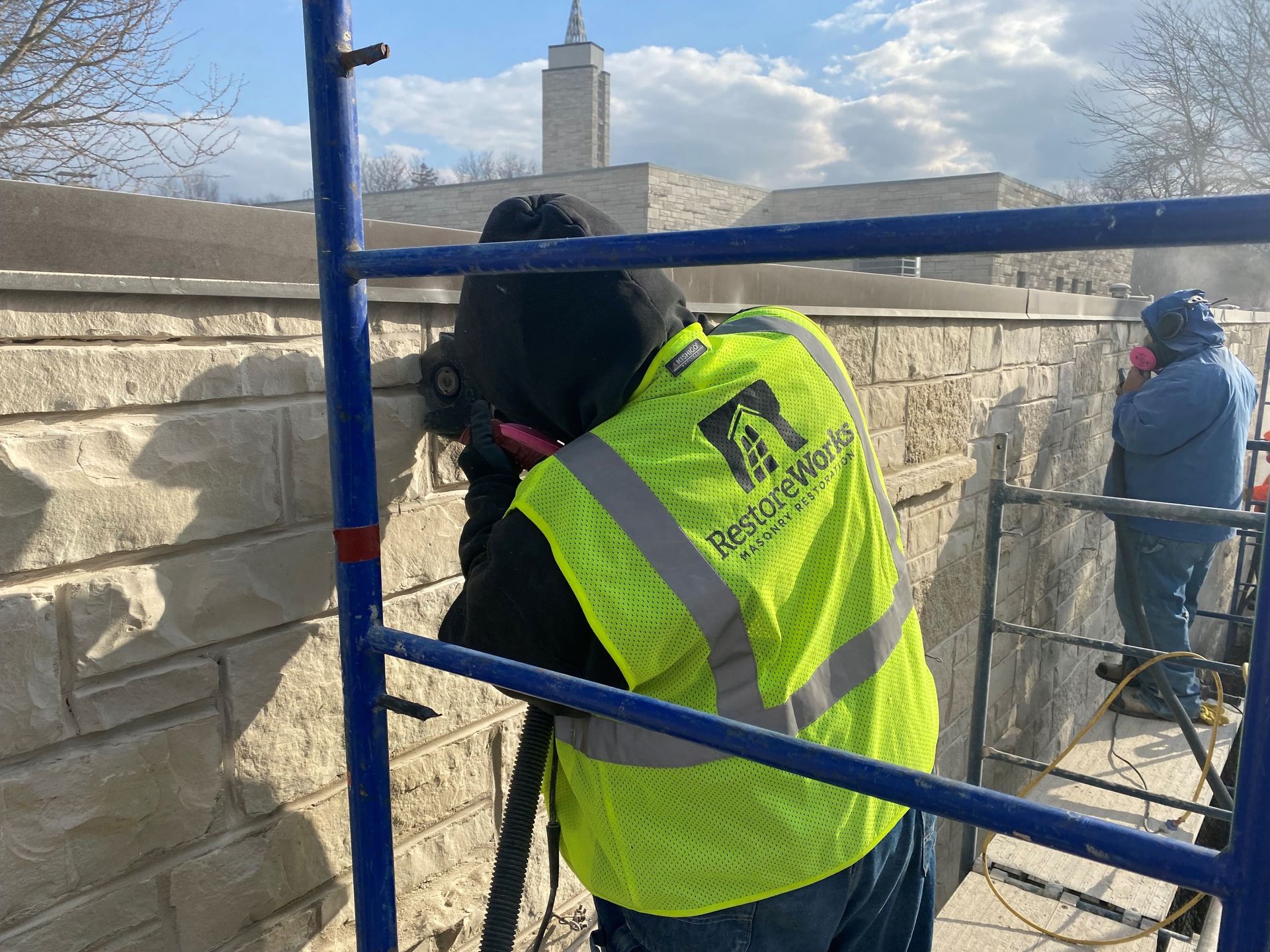 RestoreWorks masonry restoration crew performing tuckpointing on a limestone wall using power tools and dust control equipment in a commercial property setting.