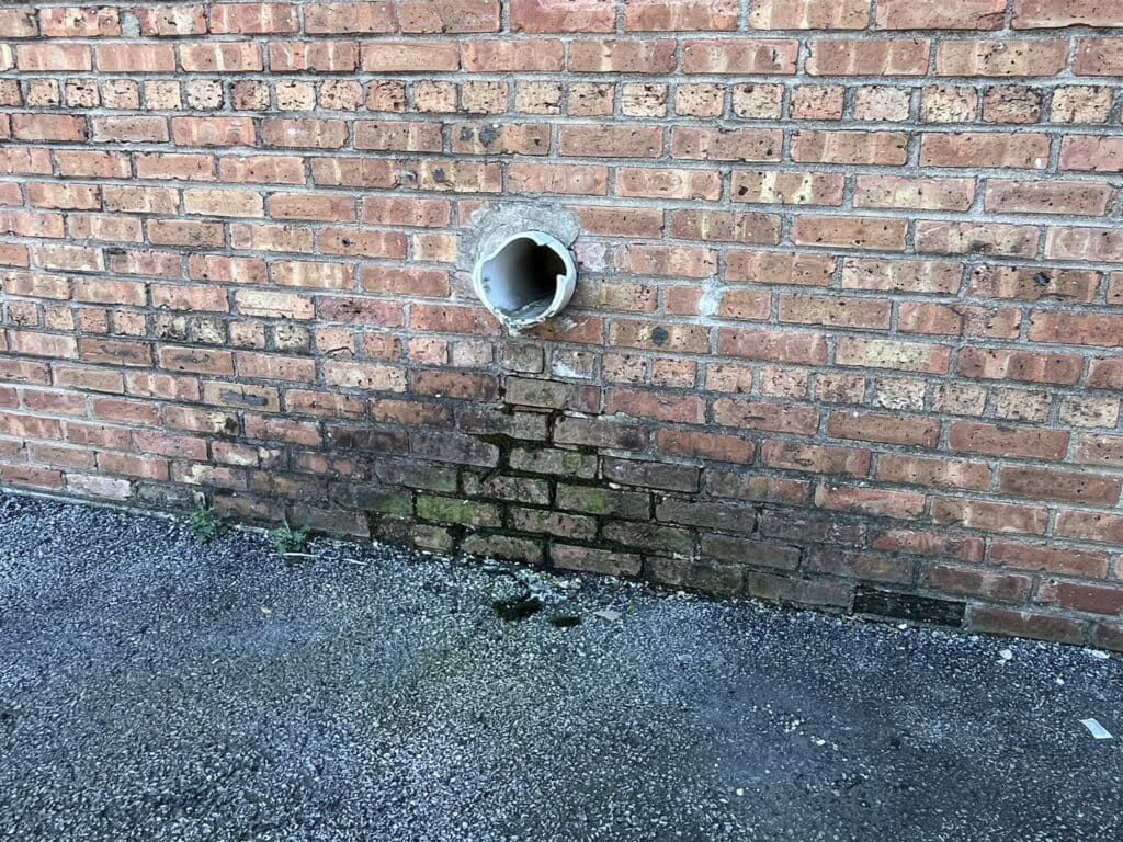 Brick wall with a round drain pipe and dark water stains on the lower bricks above gravel.