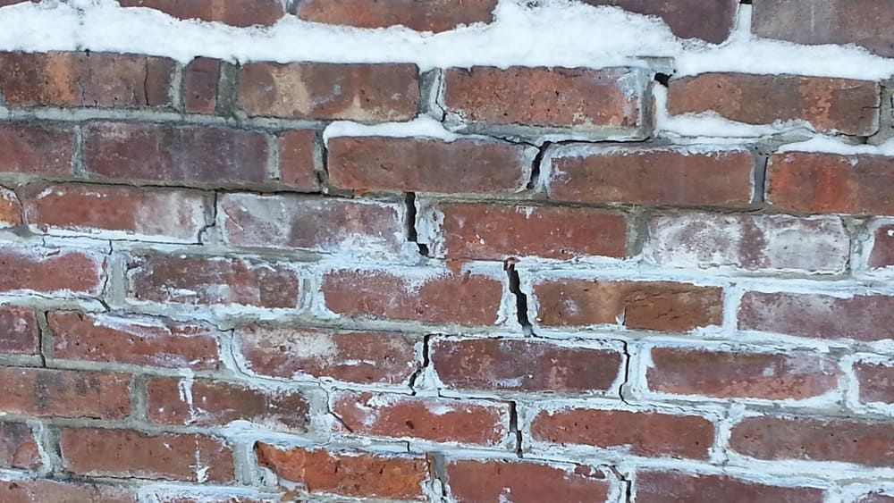 Close-up of cracked brick wall with failing mortar joints from freeze-thaw cycle damage in winter.