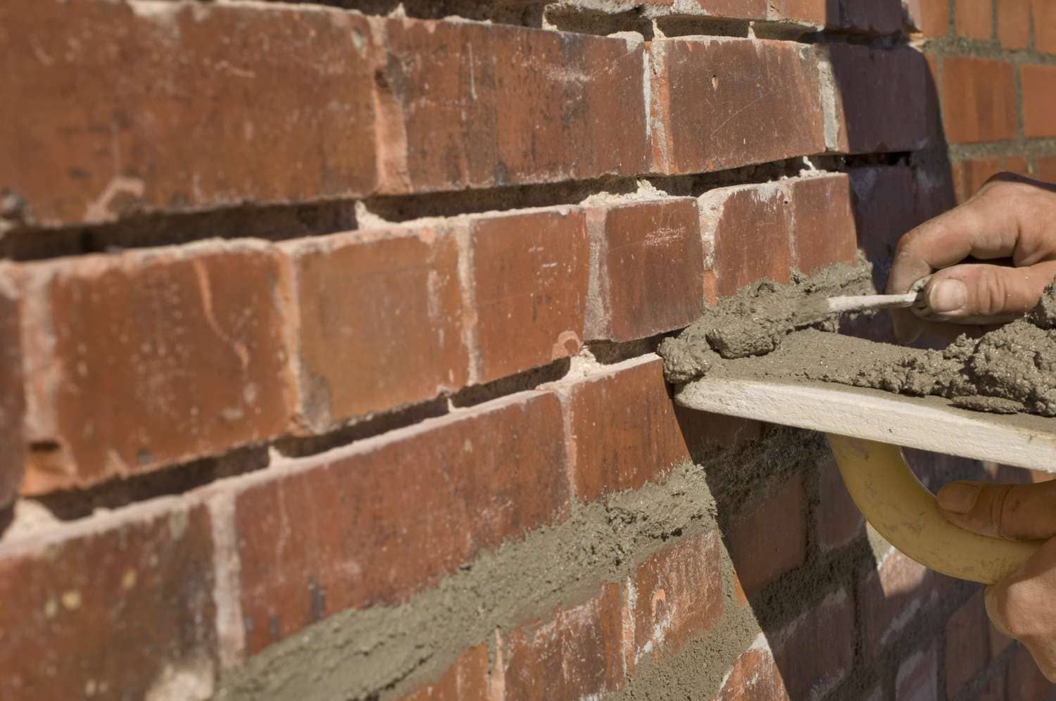 A hand uses a trowel to apply gray mortar between red bricks on a wall.