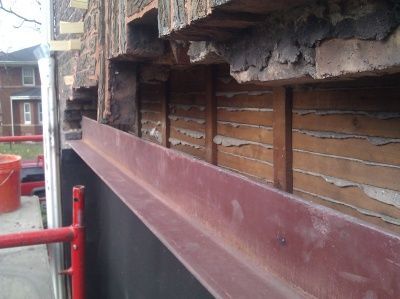 A steel I-beam is bolted to a brick building facade above wooden framing during exterior structural repairs.