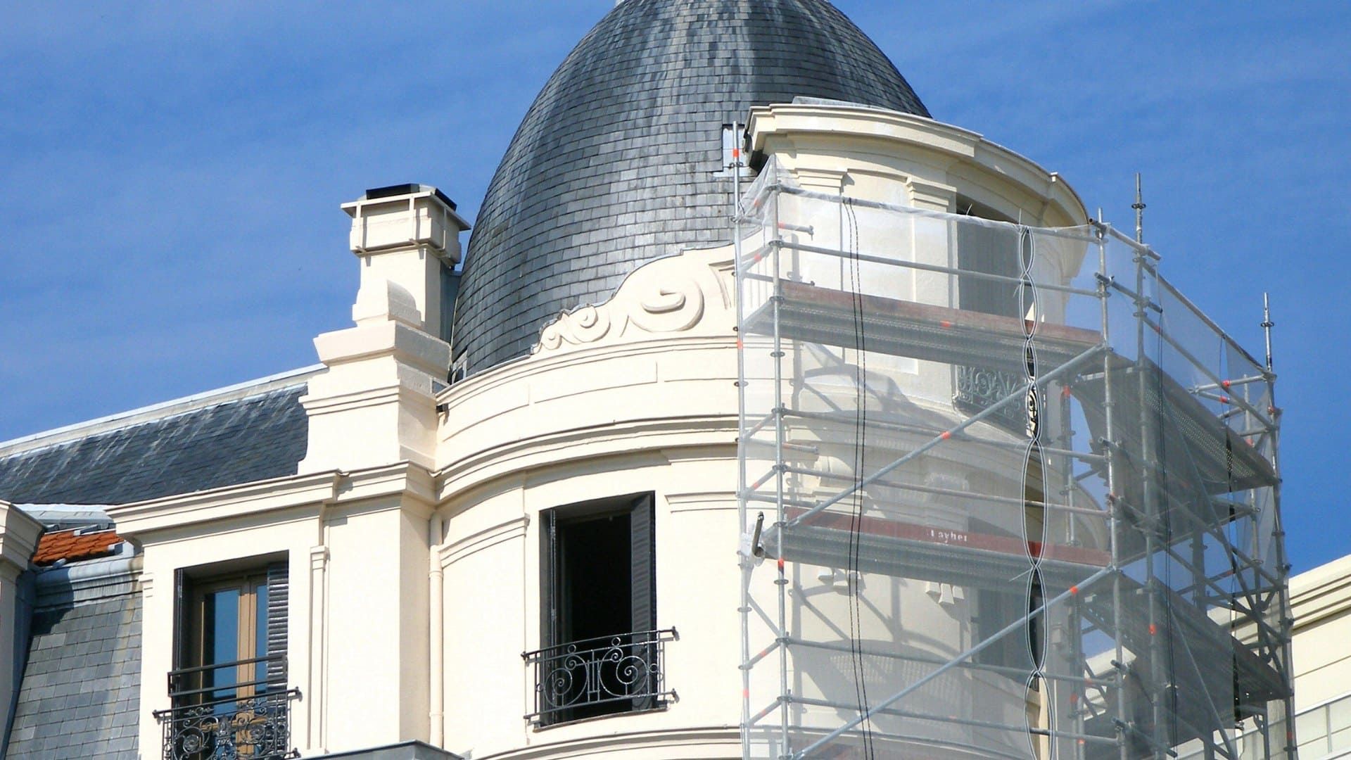 A pale building with a dark mansard roof and a round turret, partially covered by construction scaffolding.