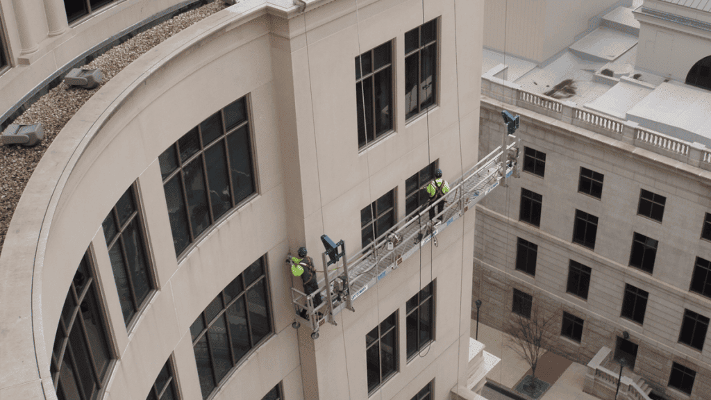 Workers on a suspended platform cleaning the facade of a tall, curved building