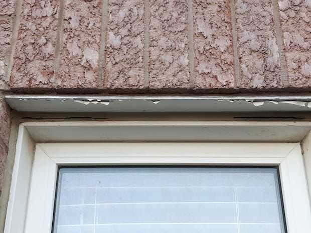 A close-up of a window frame with peeling white paint on the metal header beneath a textured, reddish-brown brick wall.