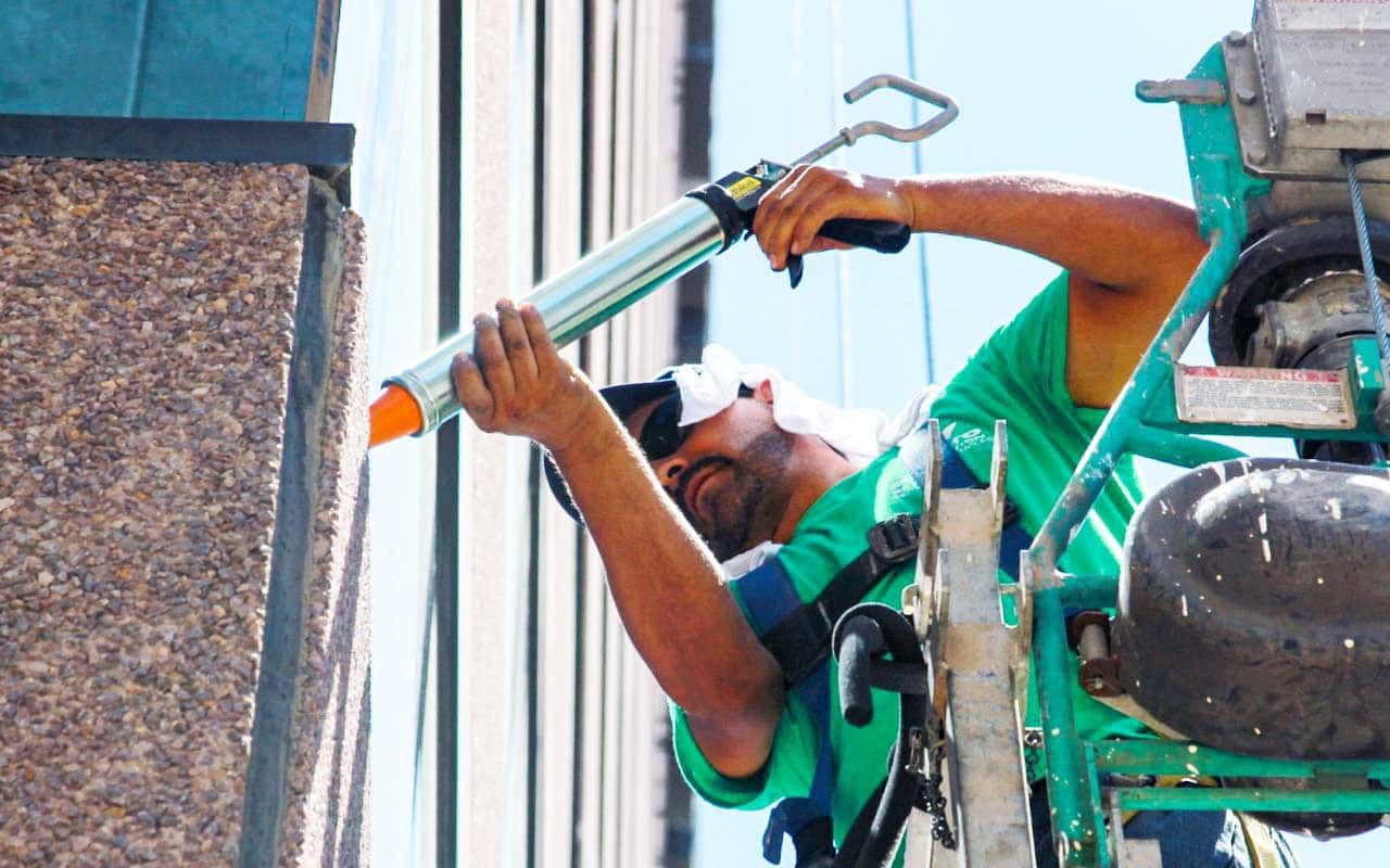 Worker in green shirt cleaning a tall window from a lift, reaching with a squeegee.