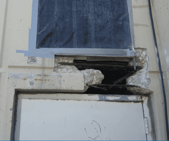 Rusted rebar exposed after concrete spalling above a doorway, showing damage that requires masonry repair and code-compliant restoration.