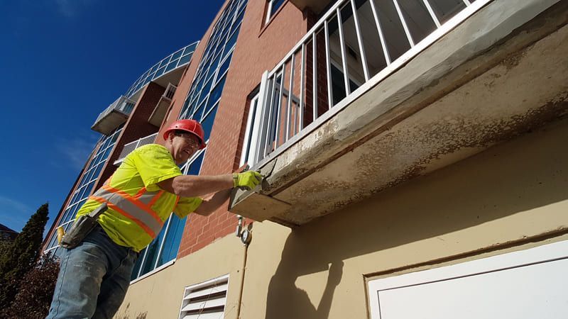 Worker in neon safety vest and red hard hat cleaning an apartment balcony exterior.