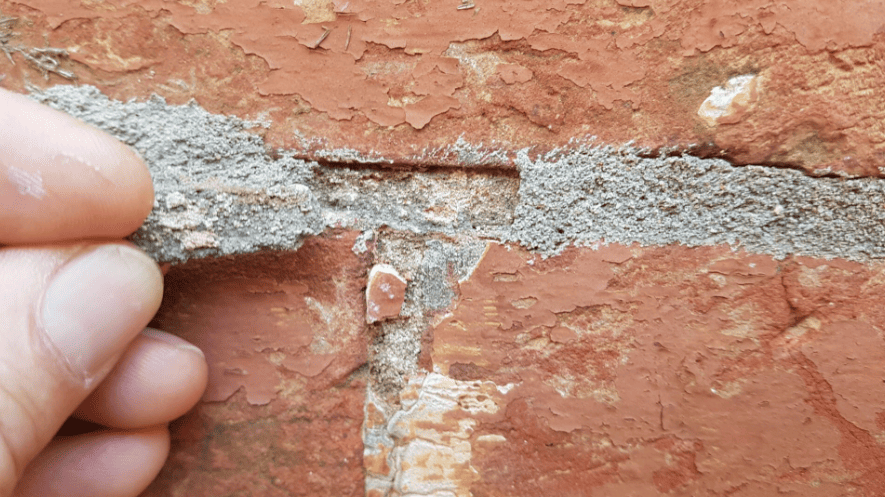 A hand peels crumbling, loose mortar away from the joints between reddish-brown bricks on a wall.