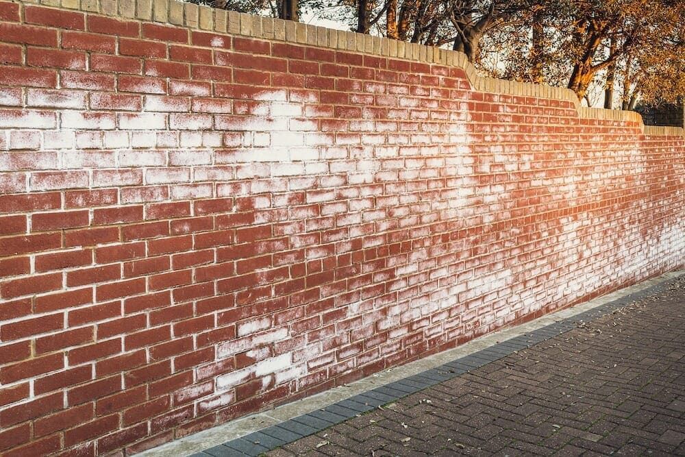 Red brick wall with white paint streaks beside a paved sidewalk and trees overhead