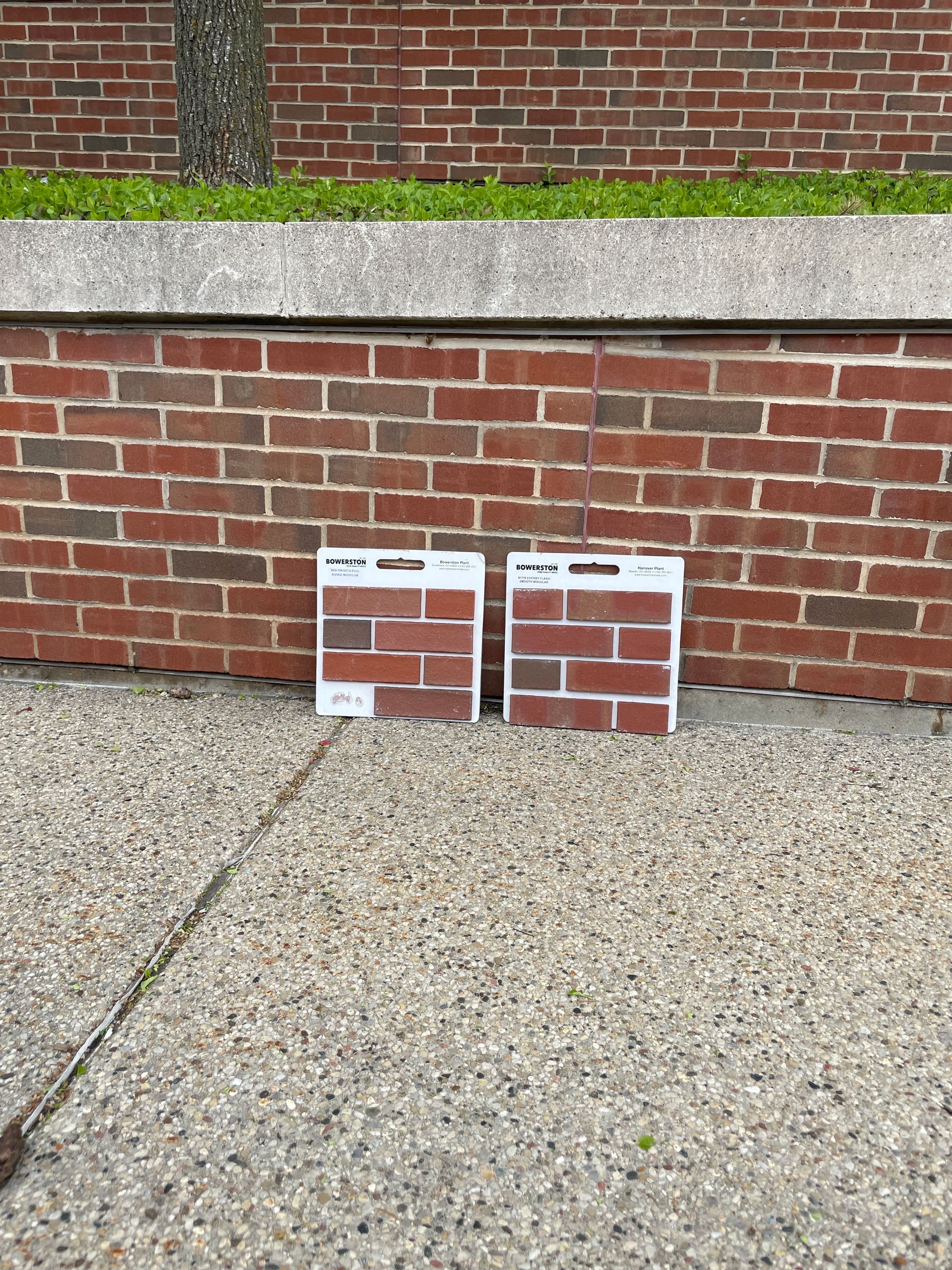 Two brick samples  are sitting on the sidewalk in front of a brick wall.