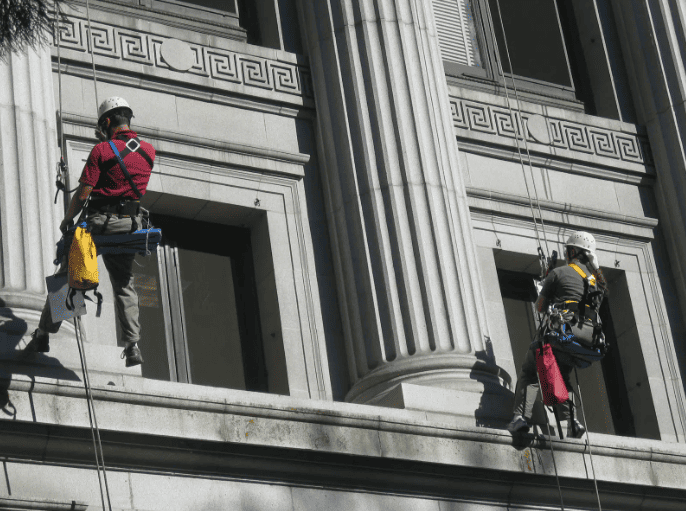 Two workers in safety harnesses suspended by ropes on the exterior of a classical stone building with large columns.