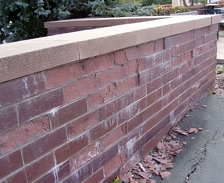 A red brick retaining wall with visible surface damage, including flaking, spalling, and white efflorescence, likely caused by freeze-thaw cycles. The wall has a concrete cap, and fallen leaves are scattered on the ground nearby. Trees and shrubs are visible in the background.