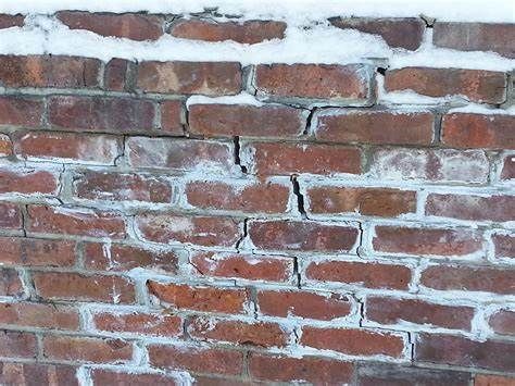 A close-up of a brick wall showing visible cracks and white efflorescence caused by freeze-thaw