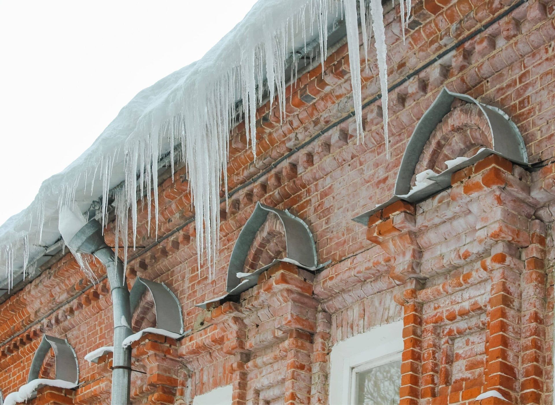 Icicles and ice buildup on clogged gutters of a historic brick building, showing poor drainage that puts pressure on masonry and increases freeze-thaw damage risk.