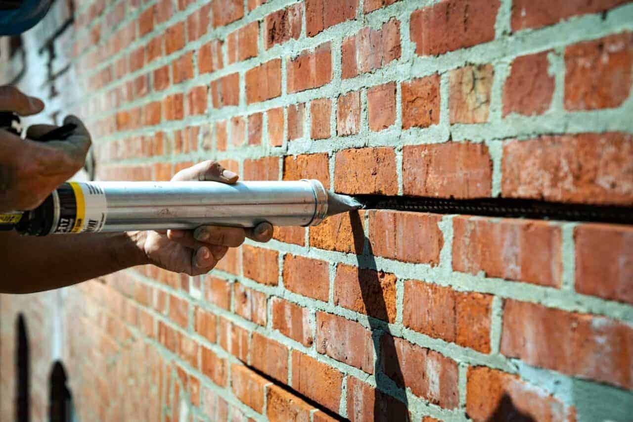 Close-up of a technician injecting grout over stainless steel helical bars installed in a horizontal bed joint, forming a hidden masonry beam to redistribute loads in a cracked brick wall during structural masonry repair.