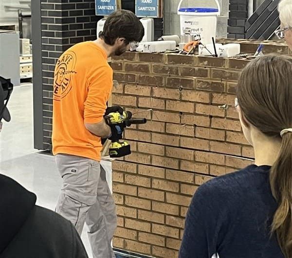Person in orange shirt using a drill on a brick wall while others watch.