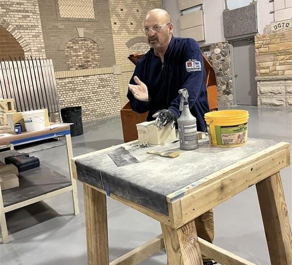 Man working at a dusty table in a stone-walled workshop, holding a tool beside paint cans and supplies