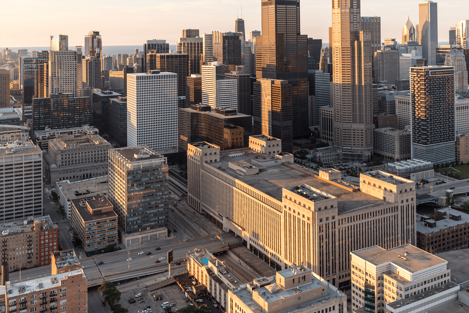 Aerial view of downtown Chicago at sunset, featuring the historic Old Chicago Post Office prominently in the foreground. The building's massive structure is surrounded by modern skyscrapers, with Lake Michigan visible in the background. The warm light highlights the architectural contrast between the historic and contemporary buildings.