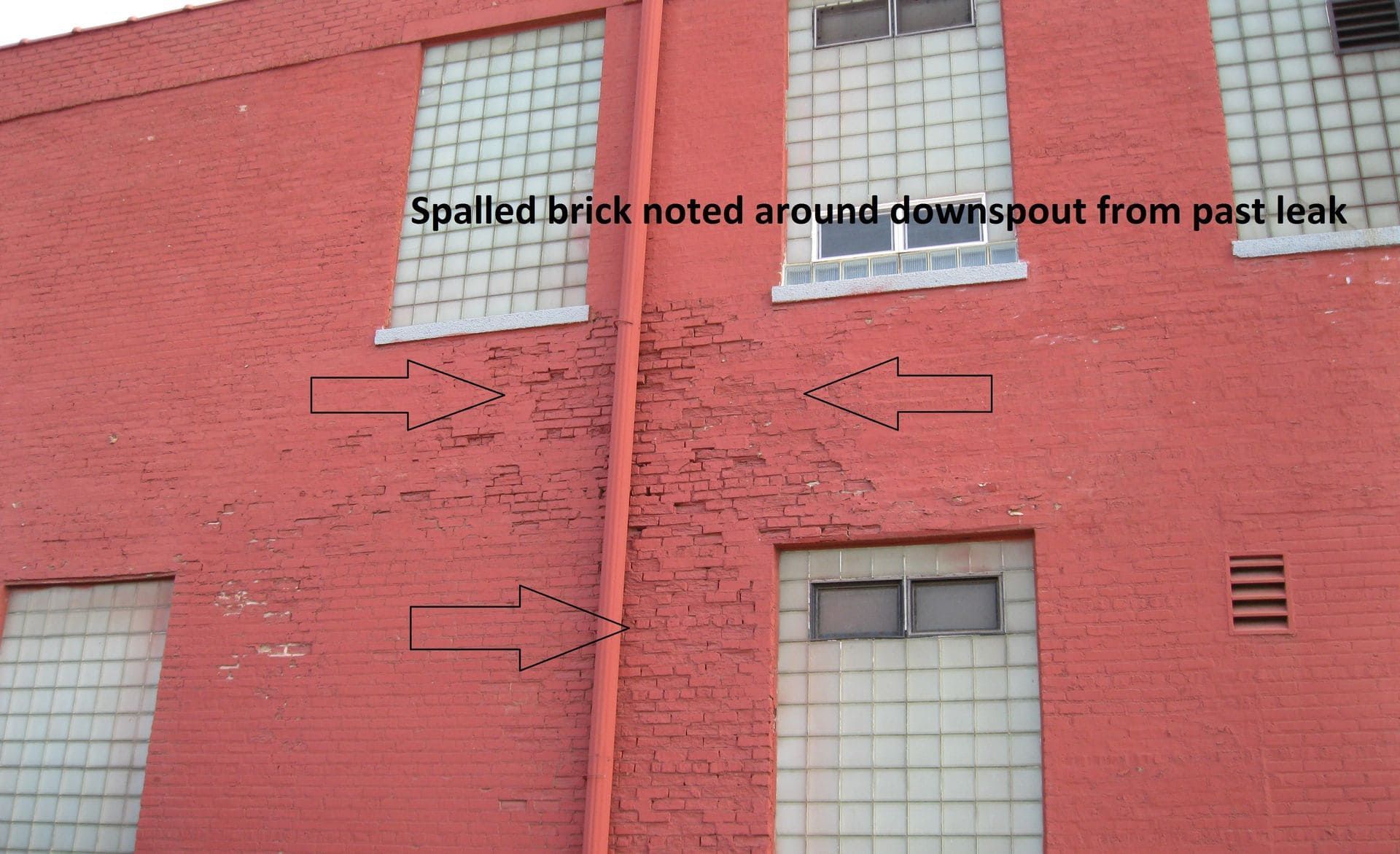 A brick building exterior showing spalled brick around a vertical red downspout, marked by arrows indicating leak damage.