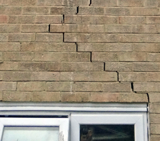 A brick wall above a window frame with a prominent, stepped diagonal crack running through the mortar and bricks.