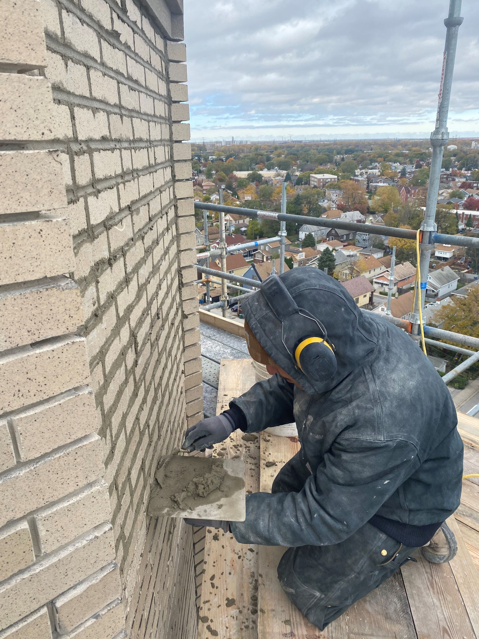 A masonry technician from RestoreWorks performing tuckpointing on a beige brick wall while working on scaffolding high above a residential neighborhood. The worker is applying mortar with a trowel, dressed in protective clothing and wearing ear protection, with a clear view of the surrounding landscape and cloudy sky in the background.