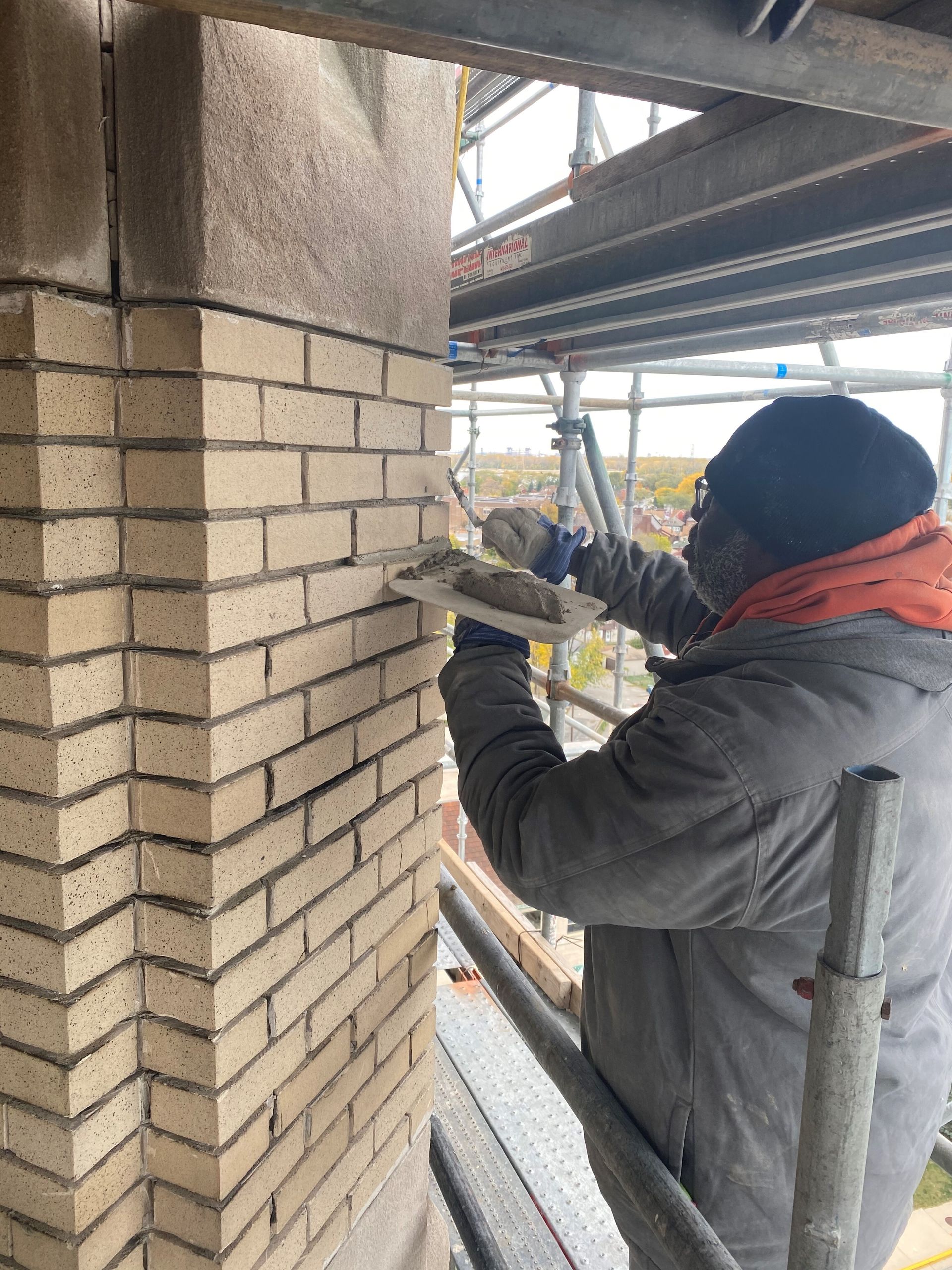 A RestoreWorks employee performing tuckpointing on a beige brick column in Northwest Indiana. The worker is on scaffolding, applying mortar with a trowel to repair and restore the brick joints. The background shows a view of the surrounding area, including trees and buildings, under overcast skies.