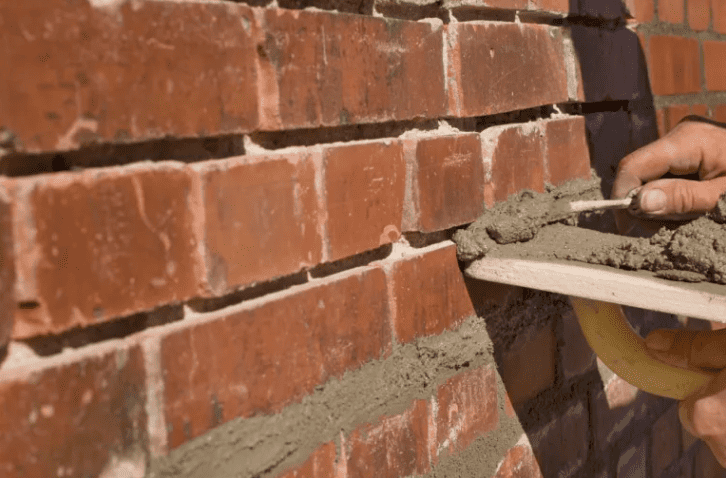 Close-up of a mason applying fresh mortar between red bricks during tuckpointing work on a deteriorated brick wall.