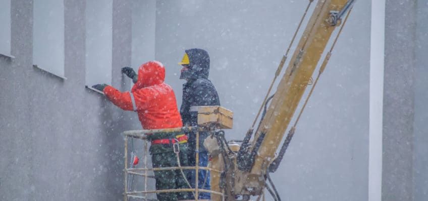 Construction crew performing concrete repair work on a building façade during snowy, cold weather conditions.