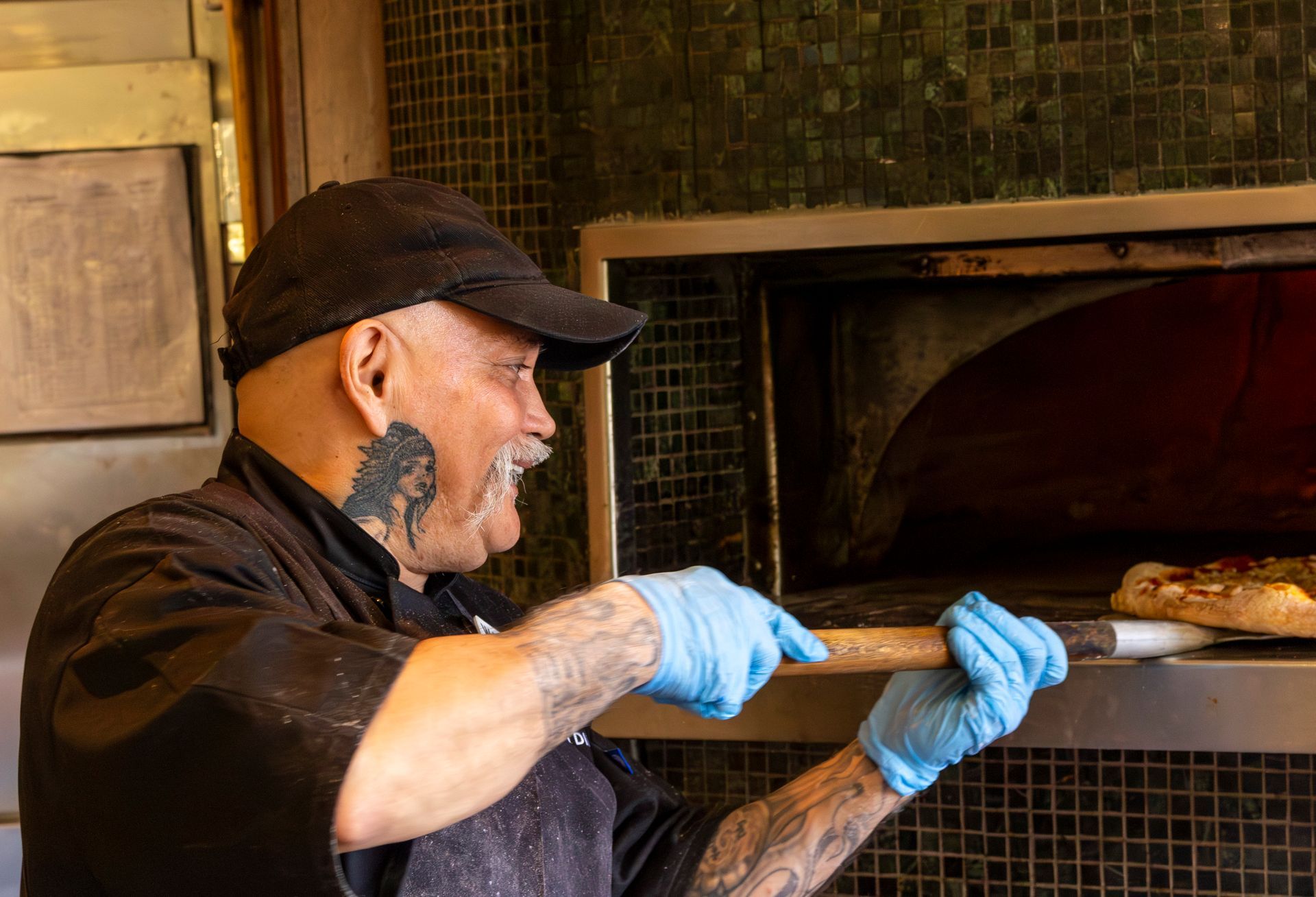 Cook wearing a black baseball cap, putting a pizza into an oven.