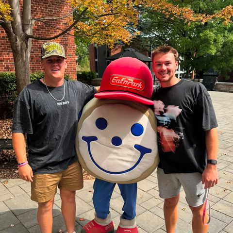 Two people stand outdoors with an Eat'n Park mascot wearing a red hat and smiling, against a brick and tree background.