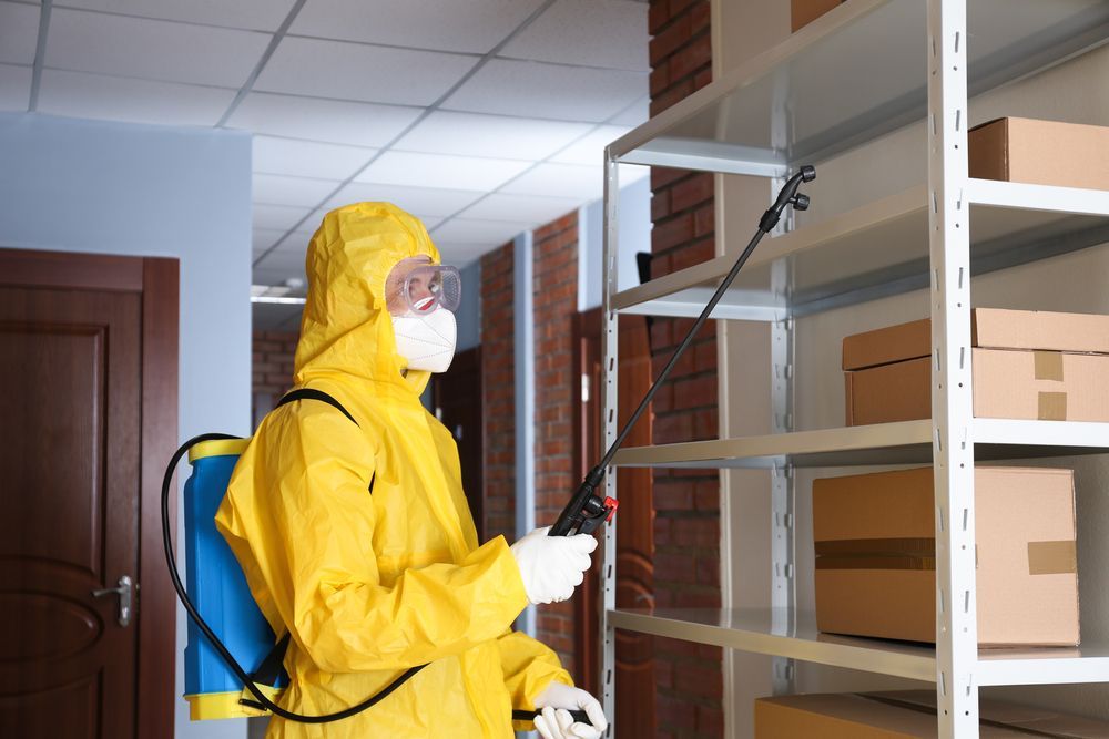 Person in yellow protective suit spraying shelves in storage room.